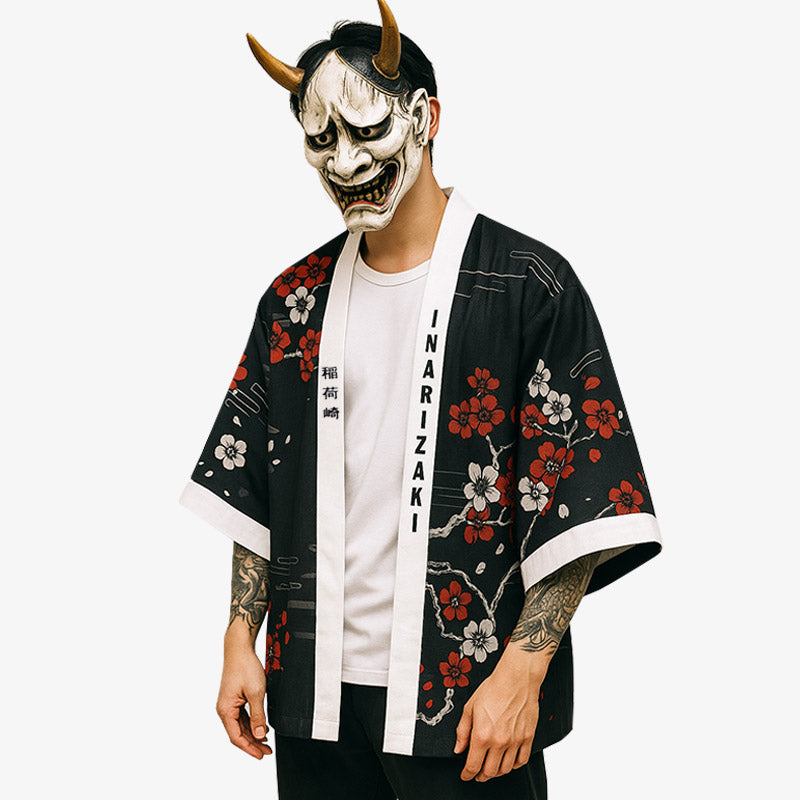 Un hombre vestido con un kimono zorro. La chaqueta haori negra tiene un estampado de flores de sakura. También lleva una máscara tradicional hannya en la cara para completar su atuendo japonés.