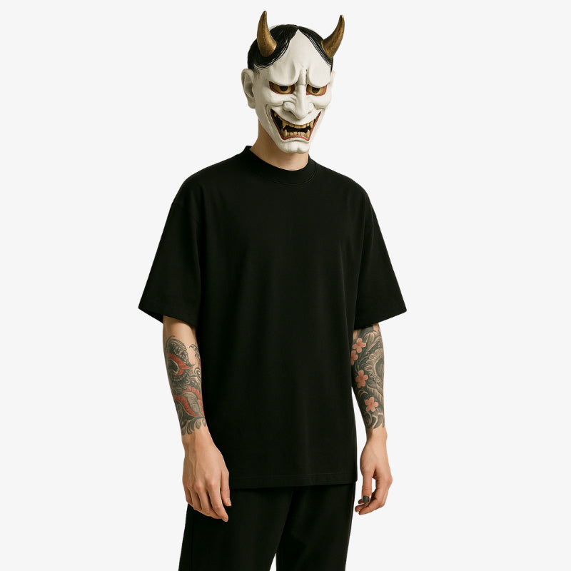 Un hombre viste una camisa estilo japones hombre totalmente negra. Su rostro está oculto tras una máscara blanca hannya. Tiene tatuajes irezumi en los brazos. La camiseta japonesa es de talla grande.