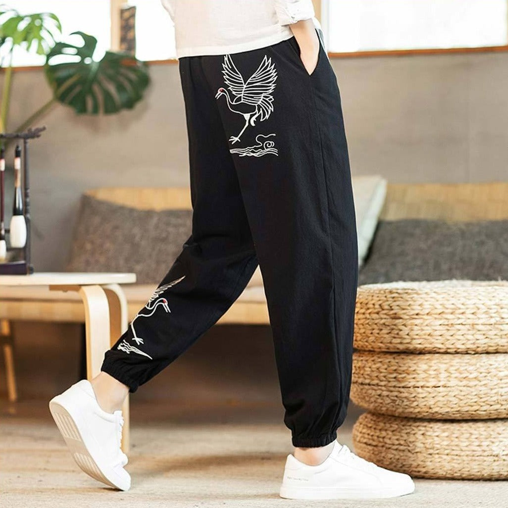 Hombre con pantalón japonés negro estampado de grullas blancas, combinado con camisa blanca y zapatillas blancas, en una habitación de suelo de madera