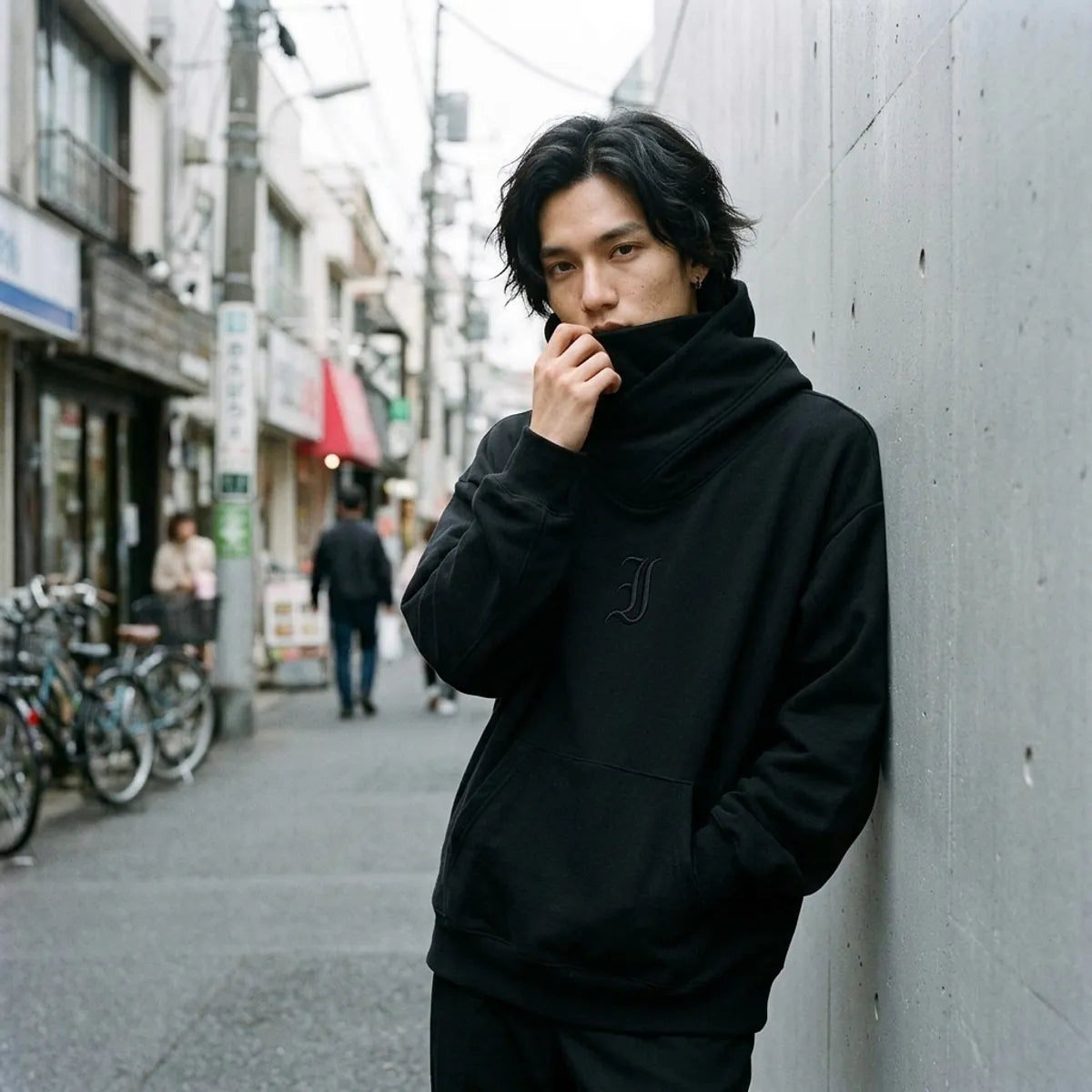 Sudadera streetwear negra de Tokio llevada con el cuello tipo chimenea subido sobre la parte baja del rostro, junto a una pared