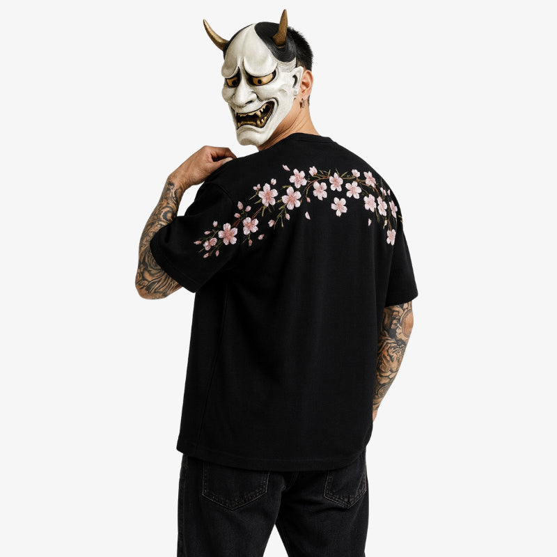 Persona con una Camiseta-Sakura negra con diseños florales japoneses, sosteniendo una máscara Hannya con cuernos y una expresión feroz.