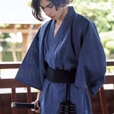 Una persona lleva un Kimono Hombre Azul azul para hombre. El kimono japonés se ata con un cinturón obi negro.