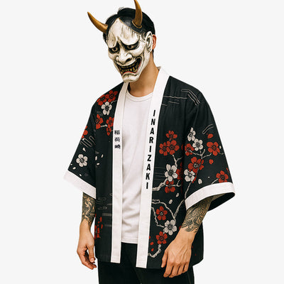 Un hombre vestido con un kimono zorro. La chaqueta haori negra tiene un estampado de flores de sakura. También lleva una máscara tradicional hannya en la cara para completar su atuendo japonés.