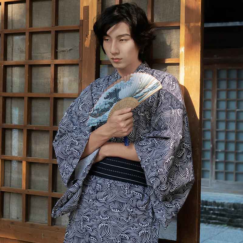 Una persona vestida con Yukata Hombre Tradicional japonés y estampado de olas. Sostiene en su mano un abanico japonés con el estampado de la gran ola de Kanagawa. El kimono se cierra con un cinturón obi negro.
