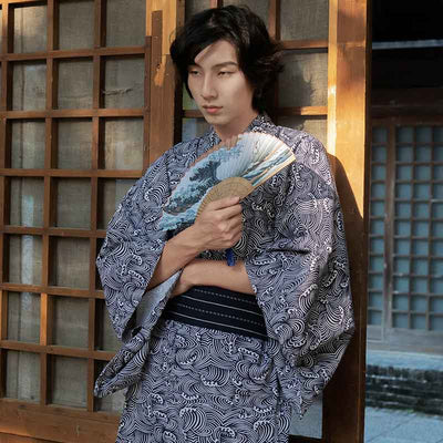 Una persona vestida con Yukata Hombre Tradicional japonés y estampado de olas. Sostiene en su mano un abanico japonés con el estampado de la gran ola de Kanagawa. El kimono se cierra con un cinturón obi negro.