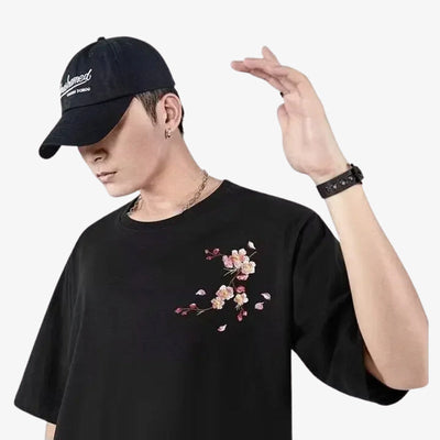 Persona con una camiseta sakura negra con diseño floral y una gorra negra sobre fondo blanco.