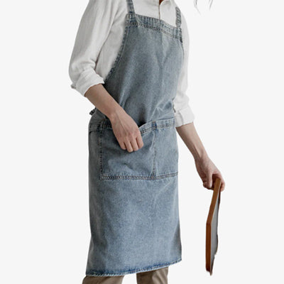 Delantal de denim confeccionado en grueso algodón azul, con tirantes cruzados en los hombros y un práctico bolsillo frontal para un uso versátil