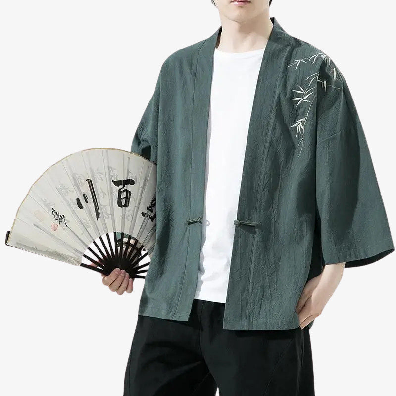 Persona con un cárdigan japonés haori verde con bordados de bambú, sosteniendo un abanico con texto kanji japonés en negro.