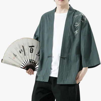 Persona con un cárdigan japonés haori verde con bordados de bambú, sosteniendo un abanico con texto kanji japonés en negro.