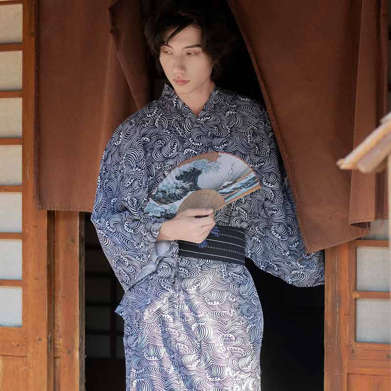Un hombre está de pie y vestido con un kimono japonés estampado con olas. Sostiene un abanico japonés sendu estampado con un patrón de la gran ola de Kanagawa del artista Hokusai.