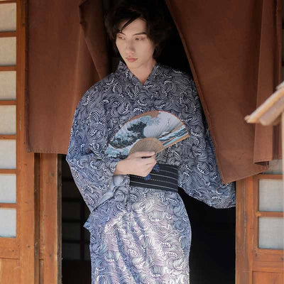 Un hombre está de pie y vestido con un kimono japonés estampado con olas. Sostiene un abanico japonés sendu estampado con un patrón de la gran ola de Kanagawa del artista Hokusai.