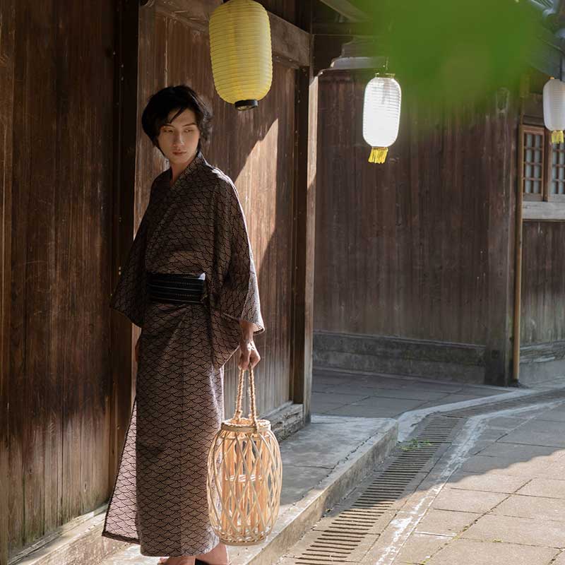 Un hombre japonés está de pie en una calle de Kioto. Viste un kimono japones seigaiha con motivos japoneses y un cinturón obi japonés. Sostiene una linterna japonesa.