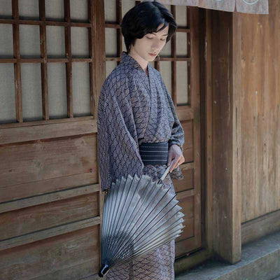 Un hombre japonés lleva un kimono seigaiha. El kimono japones es de algodón. El tejido del yukata tiene estampados motivos japoneses de olas. La prenda tradicional se ata con un cinturón obi. El hombre sostiene una sombrilla japonesa.