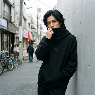Sudadera streetwear negra de Tokio llevada con el cuello tipo chimenea subido sobre la parte baja del rostro, junto a una pared