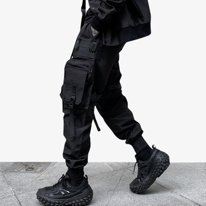 Un hombre viste techwear pantalones negros con múltiples bolsillos. Lleva zapatillas deportivas negras y una chamarra japonesa negra de estilo urbano.