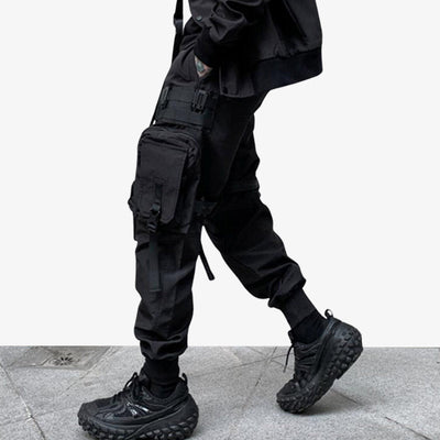 Un hombre viste techwear pantalones negros con múltiples bolsillos. Lleva zapatillas deportivas negras y una chamarra japonesa negra de estilo urbano.